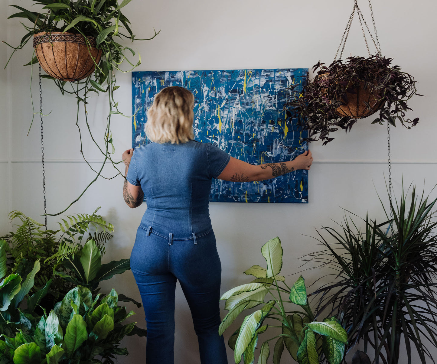 Artist hanging a large blue abstract painting on a wall surrounded by plants.
