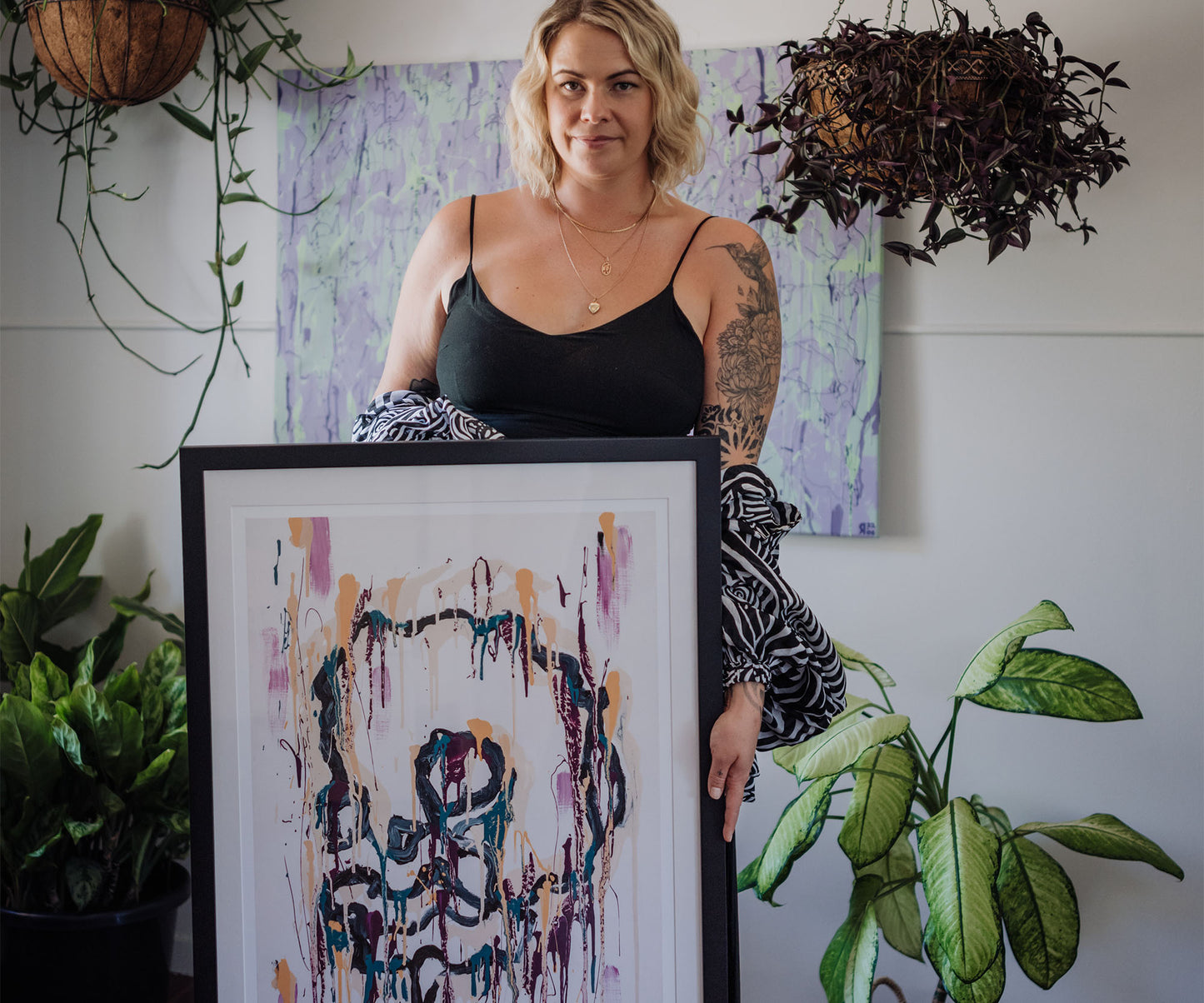 Artist Jana Liamkina holding a framed abstract art print by The Jana Saga in a plant-filled interior.