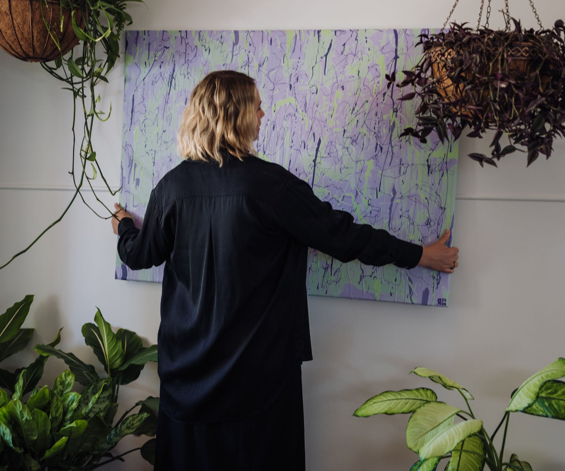 Person hanging a large purple abstract painting on a wall surrounded by indoor plants.