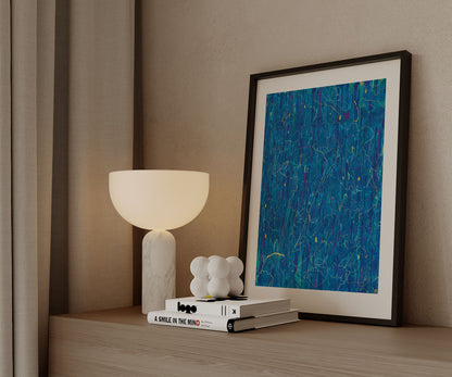 Framed blue abstract wall art print by The Jana Saga styled on a wooden cabinet beside a modern table lamp.