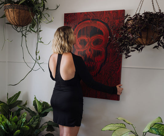 Artist hanging a large red abstract skull artwork by The Jana Saga on a wall surrounded by plants.